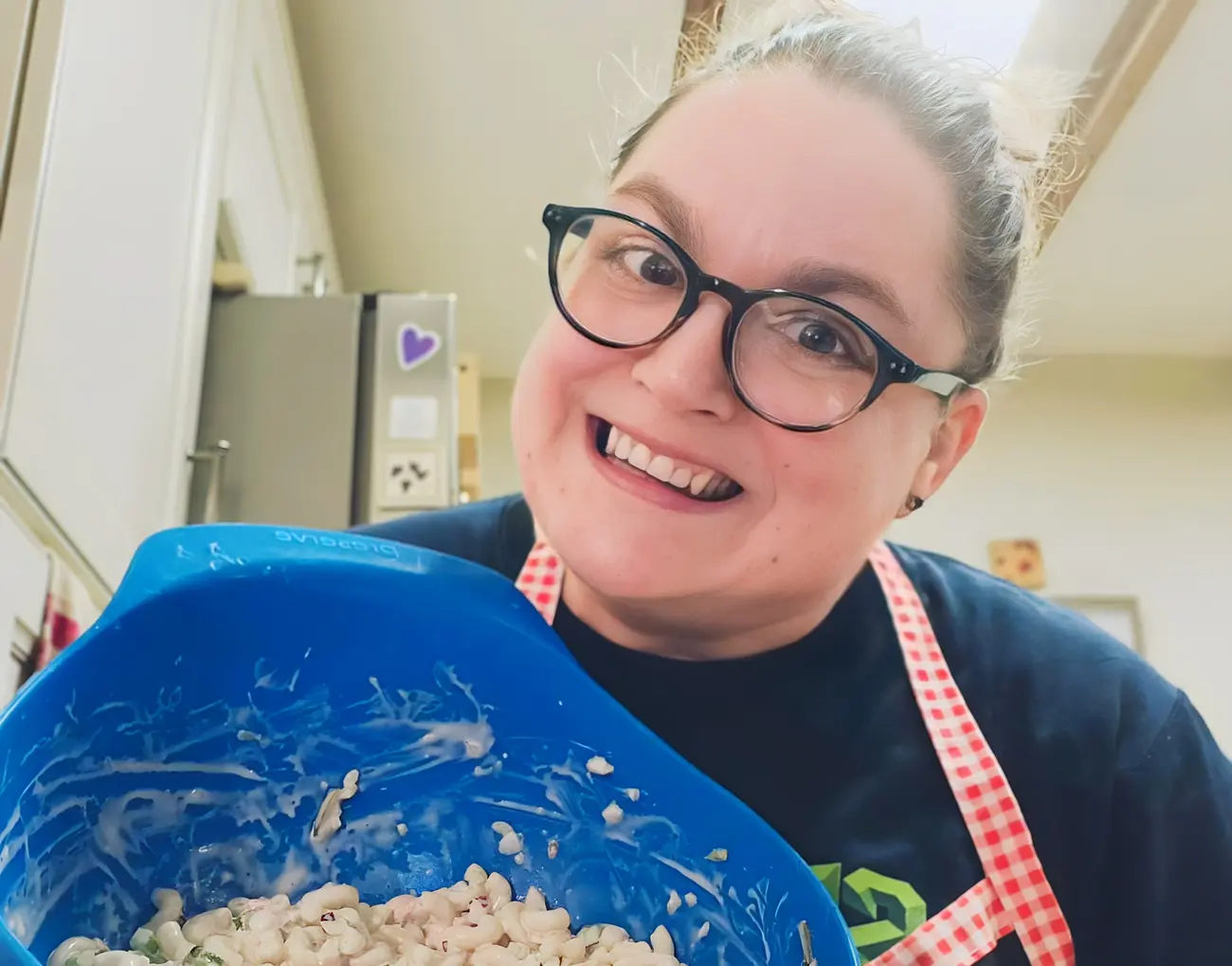 A woman with glasses and a black shirt proudly presents a bowl of macaroni salad in her kitchen, exuding joy and warmth.